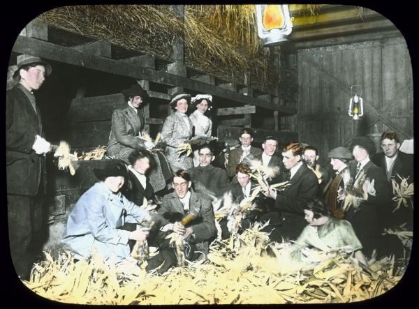 Group of men and women in a barn with piles of corn. They appear to be shucking the corn. Hay is piled in the hay mow above the group. Hand-tinted lantern slide.