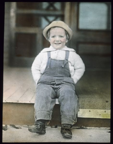 A boy wearing denim overalls, sweater, hat and shoes is sitting on a porch with his hands in his pockets. Hand-tinted lantern slide.