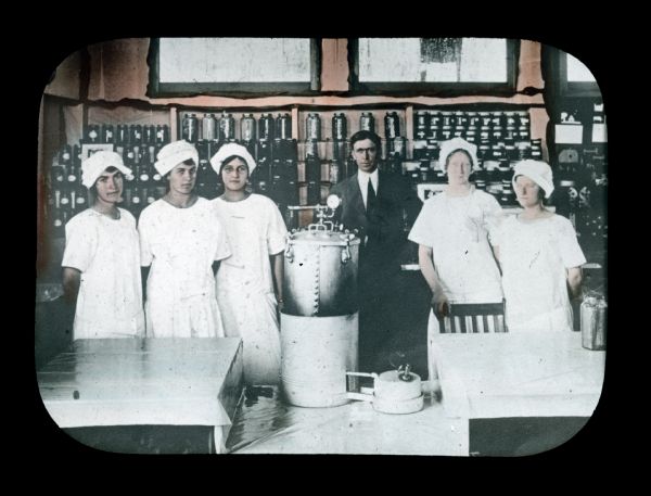 A man and five women are standing behind tables and home canning equipment. Jars of canned goods are lined up on shelves in the background. Hand-tinted lantern slide.