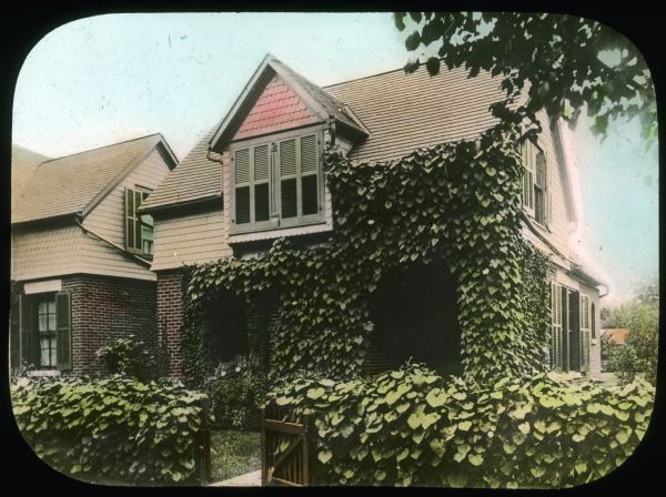 View towards front of house with vines on the fence, and vines on the side of the house over the entrance. Hand-tinted lantern slide.