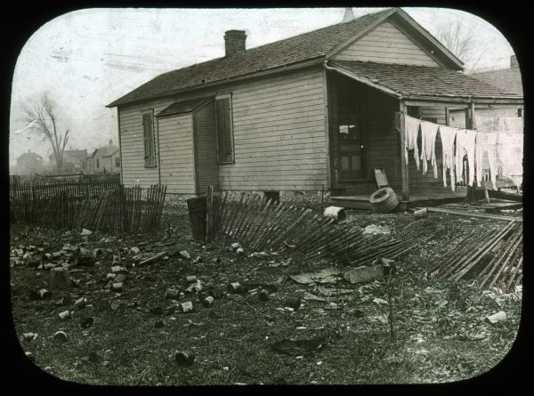 Trash-strewn lot and broken fence next to a small house. Laundry is hanging out to dry near the front of the house. Hand-tinted lantern slide.