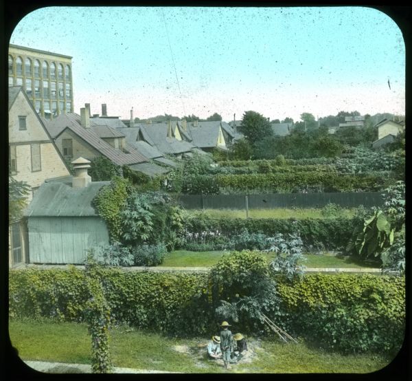 Elevated view of a number of backyards after planting. There is a factory building in the background on the left, and children playing in sand in the foreground. Hand-tinted lantern slide.