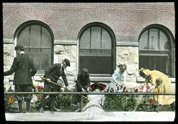 Children, boys and girls, are working in a garden at the school. The stone and brick school building has arched windows. Hand-tinted lantern slide.