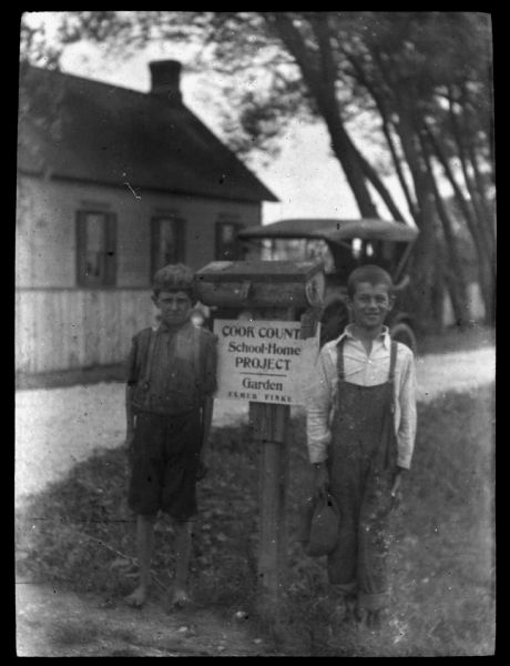 Two boys are standing next to a sign that reads: "Cook County School-Home Project, Garden, Elmer Finke. Lantern slide.