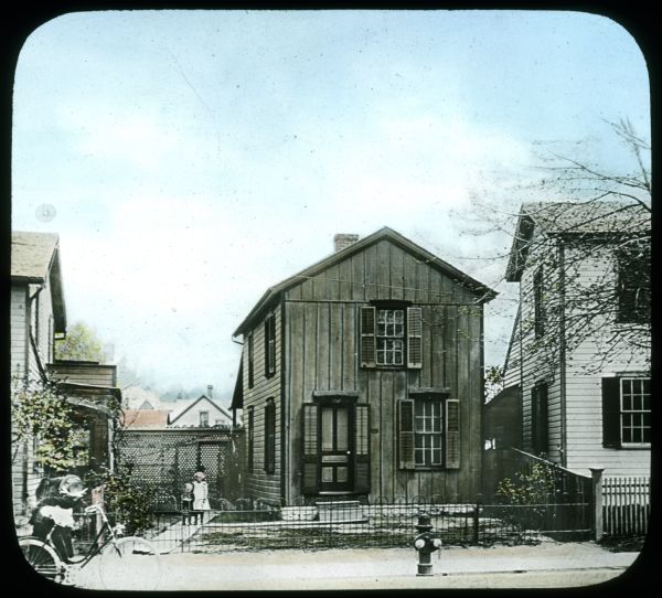 View from street towards a girl and boy standing on a walk next to a house. There is a fence in front of the house along the sidewalk. On the left is a bicycle. Hand-tinted lantern slide. Possibly the "after" photograph to follow Image ID: 137686.