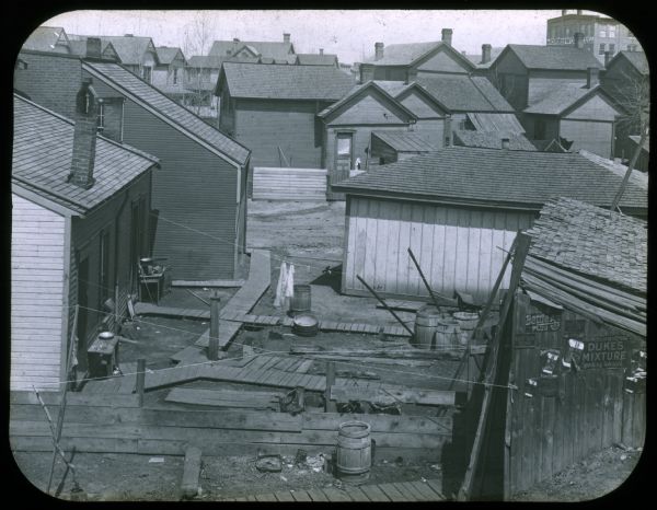 Elevated view of a backyard, with wooden walkways and barrels in an urban neighborhood. Caption reads: "Typical backyard before landscape gardening was taught." Lantern slide.