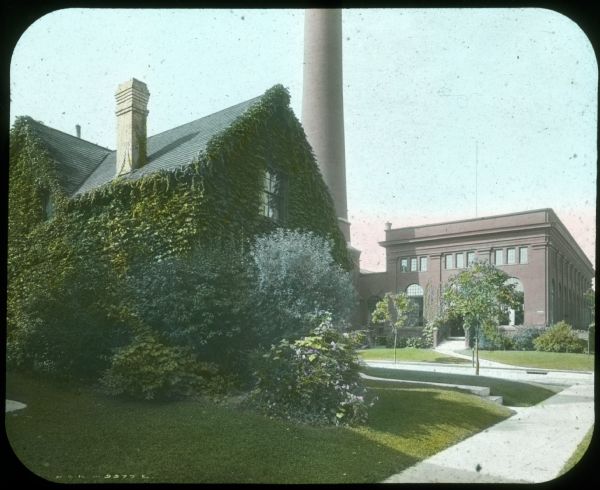House on a street corner covered with ivy and surrounded by plants and lawn. There is a factory and a large smokestack in the background. Hand-tinted lantern slide.