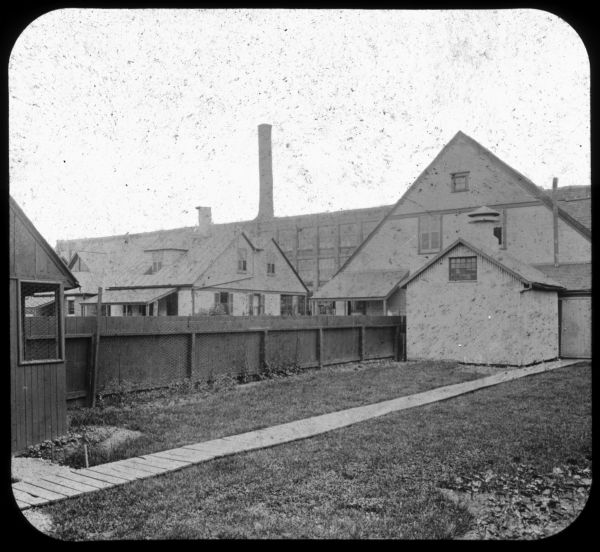 View across lawn, with wooden walkway, towards a house. A fence is on the right, and more houses are in the background on the left. In the far background is a factory building and a smokestack. Lantern slide.