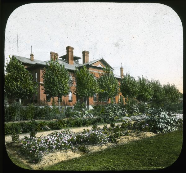 View towards a brick school building with garden in foreground. Hand-tinted lantern slide.