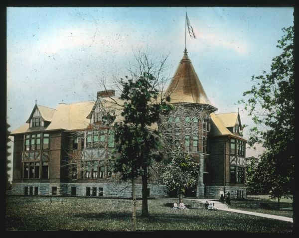 People are sitting and standing on a sidewalk and a lawn in front of a large brick and stone building. There is a turret near the entrance with a flag flying from the roof. Hand-tinted lantern slide.
