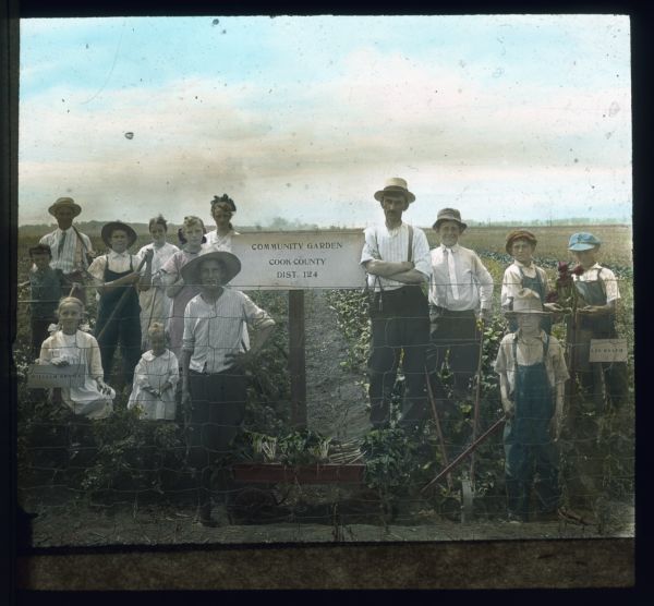 Group portrait of men, women and children standing in a field behind a fence. A sign on a post reads: "Community Garden, Cook County, Dist. 124." Hand-tinted lantern slide.