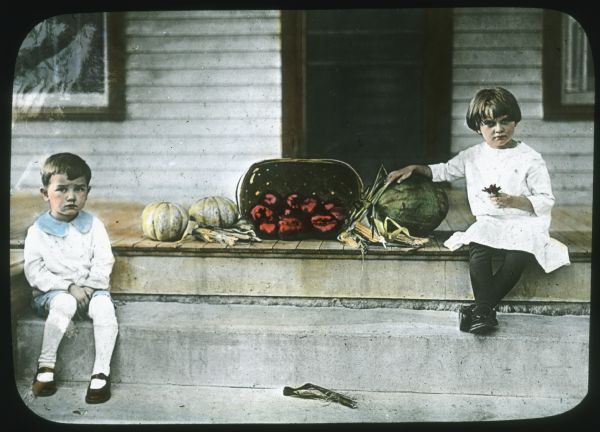A young boy and girl are sitting on porch steps of a house. On the porch is a basket of tomatoes, squash, corn, and a watermelon. Hand-tinted lantern slide.
