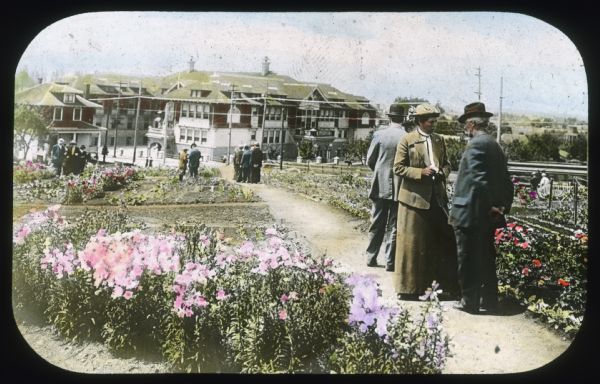 Men and women are strolling and conversing in a public garden. Hand-tinted lantern slide.