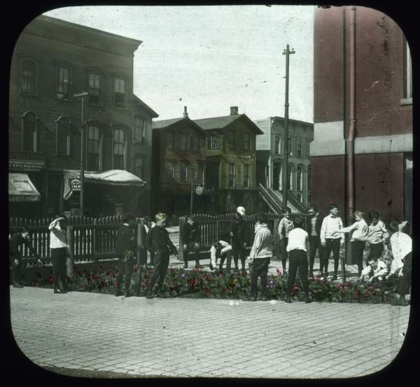 A group of boys are standing on a sidewalk around a small garden of flowers. Behind them is a fence along a city street. On the other side of the street are storefronts and houses. Hand-tinted lantern slide.
