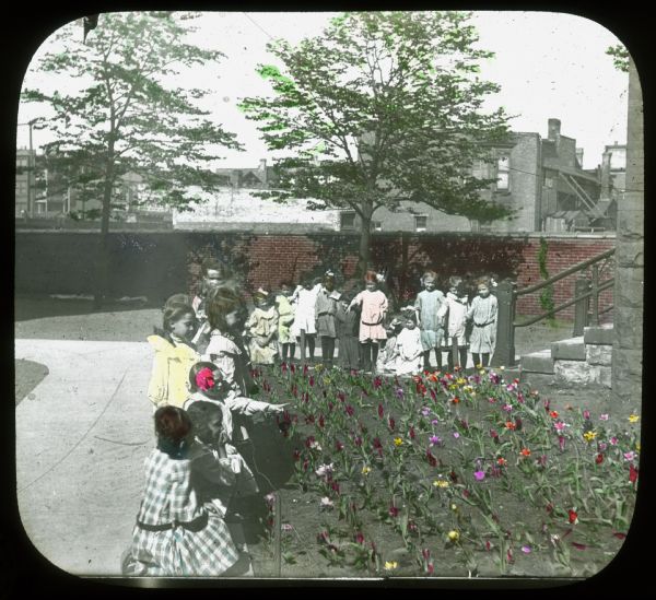 Children are standing and kneeling around a flower garden. The building on the right may be a school. There is a brick wall in the background, and beyond are houses. Hand-tinted lantern slide.