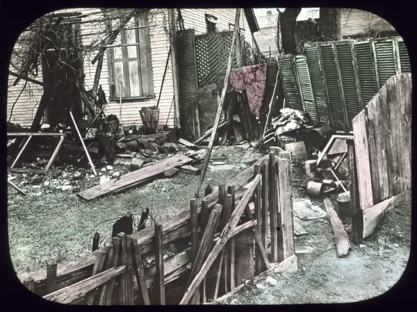 The backyard of a house is strewn with junk and flanked by broken fences. Over the back porch is a trellis with bare vines growing on it. Hand-tinted lantern slide. 