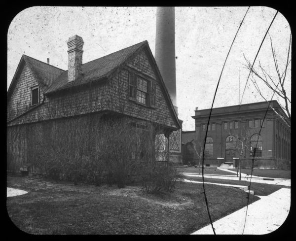 View towards lawn and shrubs around house. In the background is a factory and a smokestack. Lantern slide.