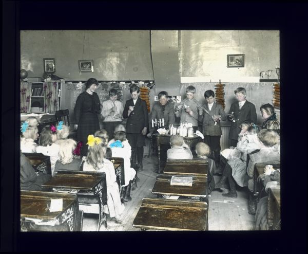 View from back of classroom towards a teacher and a group of children working with lab equipment in the front of the room. Other children are sitting in school desks at the front of the room watching. Hand-tinted lantern slide.