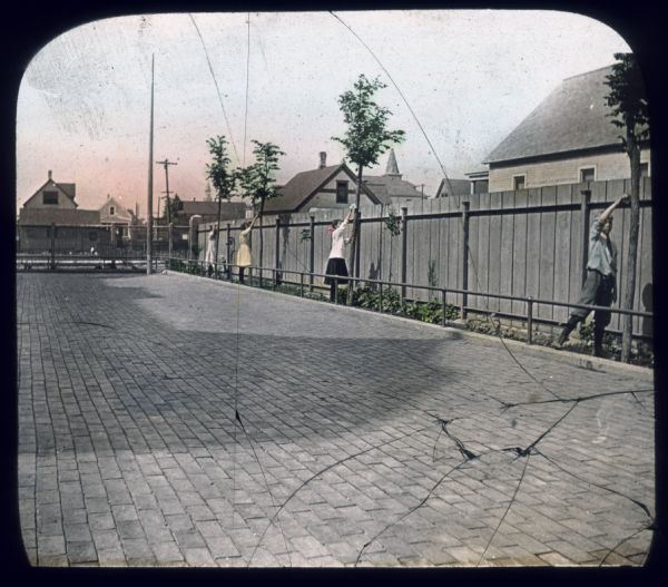 Three girls and a boy are tending to trees along a fence. A low, metal railing separates a strip of plants and trees from a brick courtyard at the "V.V. school." Hand-tinted lantern slide.