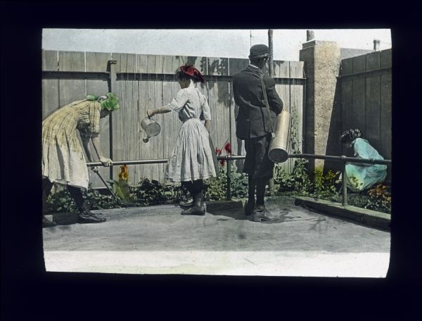 Three young women, and one young man are working in a garden in a fenced-in corner at the V.V. school. A low, metal railing separates a strip of plants and trees from a brick courtyard. Hand-tinted lantern slide.