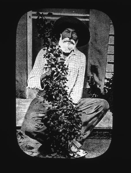 A man is holding a plant which may have berries, while sitting on a step outdoors in front of the door of a building. Lantern slide.