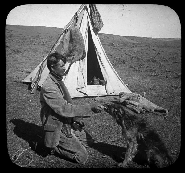 A man is kneeling on the ground and holding onto something that his dog is holding with its mouth. Behind them is a tent, with a firearm leaning against a tree limb fixed in the ground that is being used to hold up the tent. Caption reads: "13650 — The Lone Shepherd and His Best Friend, Solitudes of a Sheep Ranch, Montana, U.S.A." Lantern slide.