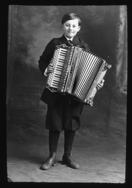 Full-length portrait in front of a painted backdrop of a smiling boy posing while holding an accordion. The name on the accordion reads: "Serenelli-Ruatta, Chicago, Illinois."  Lantern slide.