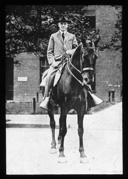 A man wearing a suit, necktie and hat is sitting on a horse. In the background is a large, brick building. Lantern slide.