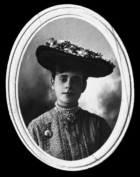 Quarter-length portrait of a woman. She is wearing a lace collar with a choker necklace, a pocket watch pinned on her dress, and a straw hat decorated with flowers. Lantern slide.