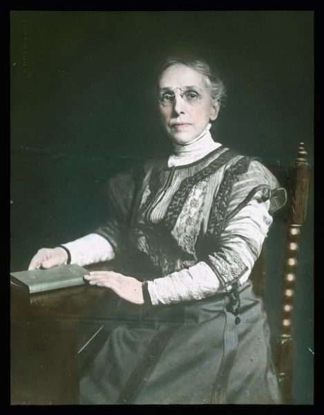 Three-quarter length portrait of a woman sitting in a chair at a table. She is holding a book on the table, and is wearing pince-nez eyeglasses.