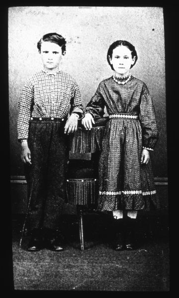 Full-length studio portrait of a boy and girl standing with their hands resting on the back of a tasseled chair. Lantern slide.