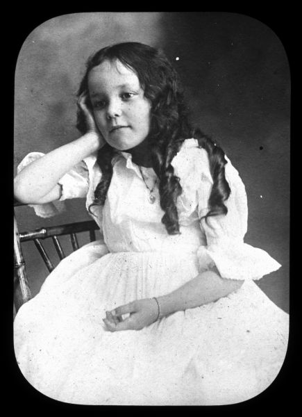 Portrait of a young girl with long curls sitting on a chair. She is leaning her right arm on the back of the chair is and is resting her head on her hand. Lantern slide.