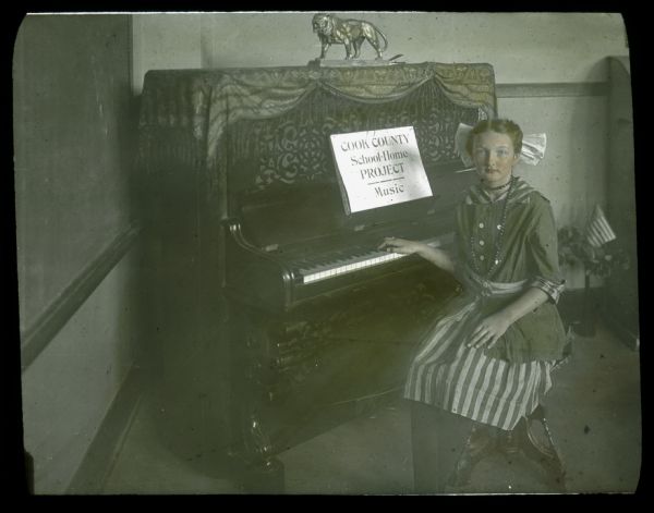 A girl is sitting at a piano. A sign on the piano reads: "Cook County School-Home Project — Music." Hand-tinted lantern slide.