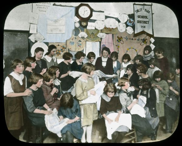 A group of girls and a woman are standing and sitting together in a classroom working with fabric, in the Tinley Park School District." Hand-tinted lantern slide.