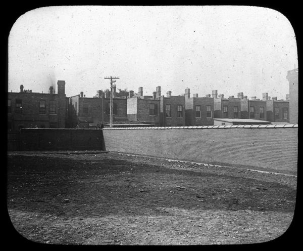 Corner of a Chicago schoolyard. There is a brick wall at the border of the yard, and brick buildings are across the street. Lantern slide.