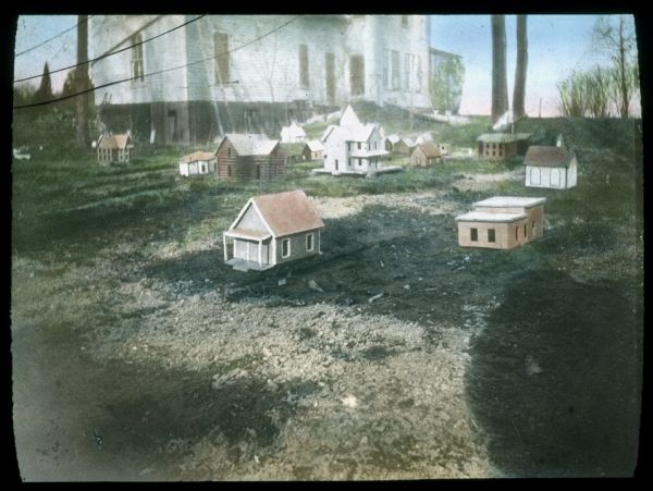 Group of small-scale model houses on the lawn in a school yard. Hand-tinted lantern slide.