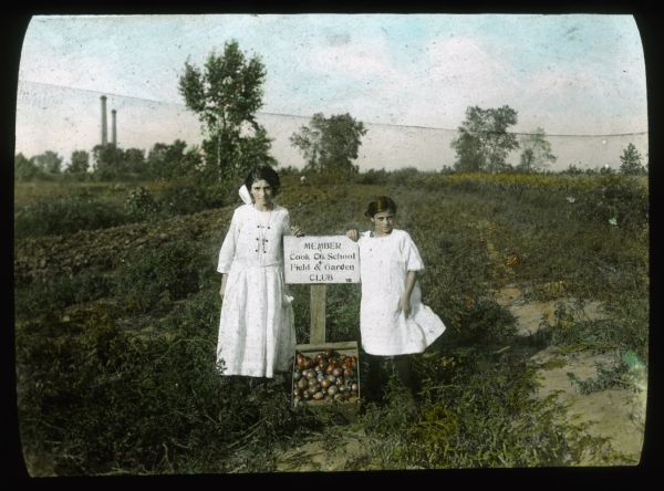 Two girls, each wearing white dresses, are posing outdoors in a garden. At their feet is a box of produce (tomatoes?), and they are leaning on a sign which reads: "Cook Co. School, Field & Garden — Club." Hand-tinted lantern slide.