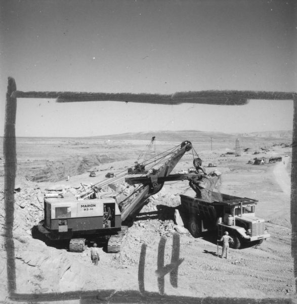 Elevated view of a crane loading an International Payhauler for earth removal. One man is standing near the crane on the left, and another man is standing near the truck on the right. Crop marks and "IH" have been drawn on the image. Caption reads: "Big Dipper for Big Ditch. Tons of desert soil must be carted from site in Payhaulers in preparation for roads and 1,028-foot-long bridge which will span canyon 850 feet below the damsite."