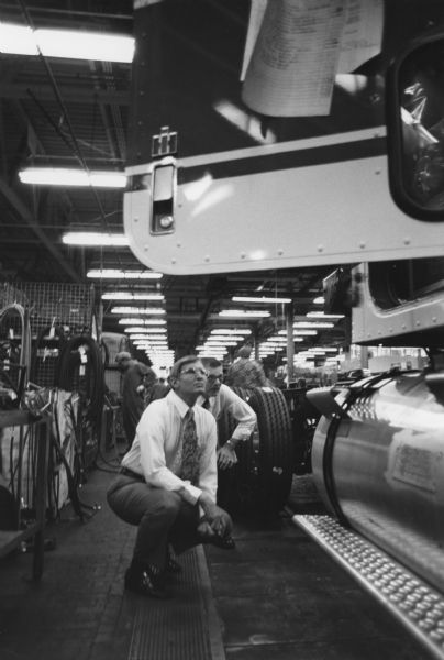 Two men are crouching on the factory floor looking up at a truck. Men are standing in the background.