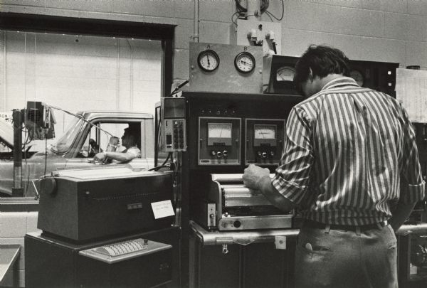 A man is in a room with testing equipment. In the background is a man sitting in the driver's seat of a truck.