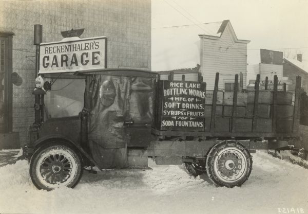 International truck owned by the Rice Lake Bottling Works parked on a snow-covered street in front of Reckenthaler's Garage. The truck is equipped with chains on the tires and a tarp over the open side of the truck. The bottling company manufactured soft drinks, syrups and fruits for soda fountains.