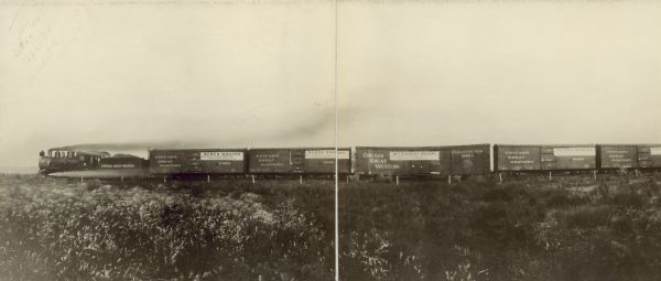 A panoramic view of the Chicago Great Western train traveling across the countryside carrying assorted International Harvester wagons. The majority of the cars contain Weber Wagons, with one car holding Bettendorf Wagons.