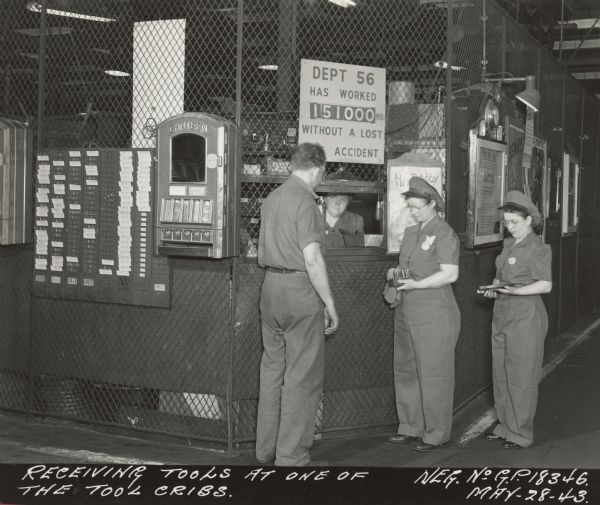 Man and two women receiving tools at tool crib. A woman is sitting inside the window of the tool crib. Also pictured are timecards, and a "Canteen" vending machine filled with "Chuckles," "Jumbo" and other snacks. A sign on the vending machine reads, in part: "Rationing has limited our supplies.". A sign on the fencing around the tool crib reads: "Dept 56 has Worked 151000 RS Without a Lost (obsucred word) Accident."