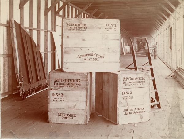 View of a McCormick Little 4 mower and two Folding Daisy reapers boxed and ready for shipment from IHC's McCormick Works. The boxes are stamped with destinations. A man is standing in the background on the right.