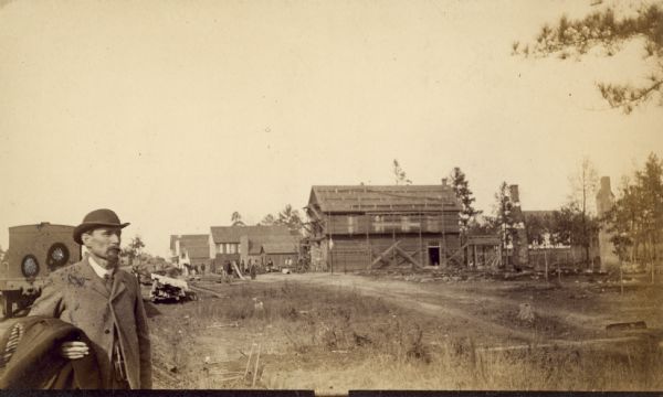A man wearing a hat and carrying a coat over his arm is standing in the left foreground. Beyond is a town, with a building under construction. Caption reads: "Catterus (unsure of spelling) New Store."