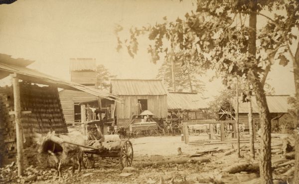 View of the cotton gin. A man is driving a small cart in the left foreground. Men are standing in front of cotton gin buildings in the background.