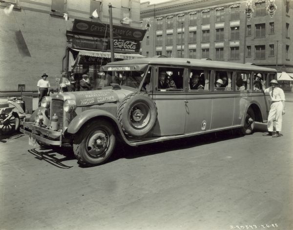 Group of men, likely World War I veterans, sitting in an International model 52 or model 53 bus in the streets of Omaha. One man is standing outside near the back of the bus, and another man is looking on from the sidewalk. The bus is parked near the "Crystal Candy Co." storefront, and is decorated with a banner on the front hood which reads: "International Harvester Motor Coaches." Three flags are mounted on the hood ornament, one of which may be the British union jack.