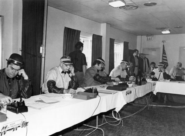 Salesmen are sitting at tables talking on telephones. Caption reads: "Here's the International 'Blitz A Grand' salesmen in action. (From left) John Stills, Harry Garrison, Bob Armstrong, Bill Hough and Dale Zimmerly donned derbies, sleevelets and eyeshades to create an old-time 'boiler room' atmosphere while attempting to sell 1,000 all-wheel drive pickup and Loadstar medium-duty models in a single day by telephone to International dealers in Missouri, Kansas, Iowa and Nebraska. The results far exceeded any predictions as a total of 1,330 4x4 pickups and 410 Loadstar units were sold by 15 International key market managers and zone managers in an eight-hour period April 26."