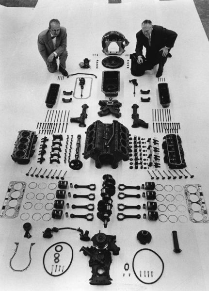 Elevated view of two men who are posing with the parts of a V8 engine displayed on the floor on top of a white background.