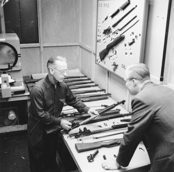 Elevated view of two men in a room standing at a table with rows of rifles on it. One of the men is holding one of the partially disassembled rifles. Above the table on the wall is a framed board that reads: "U.S. Rifle" and the disassembled parts of the rifle displayed on it.